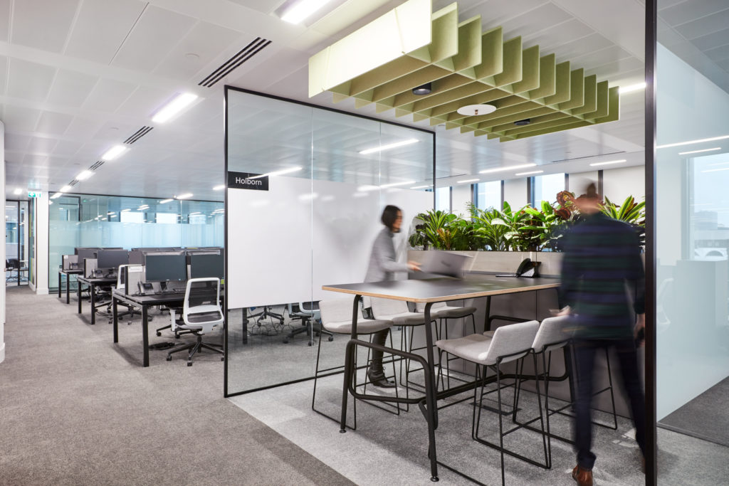 Open plan office space with desk area and glass divider separating high table and stools. 