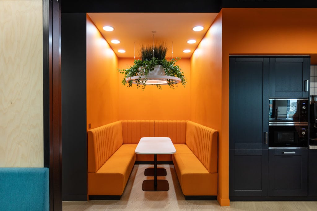 Workplace refurbishment with orange seating booth with hanging plant light feature next to office kitchen area.