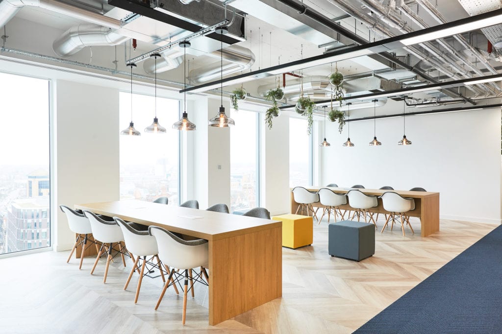 Gestaltung eines kollaborativen Büroraums mit zwei großen Tischen, Beleuchtungselementen und Hängepflanzen.