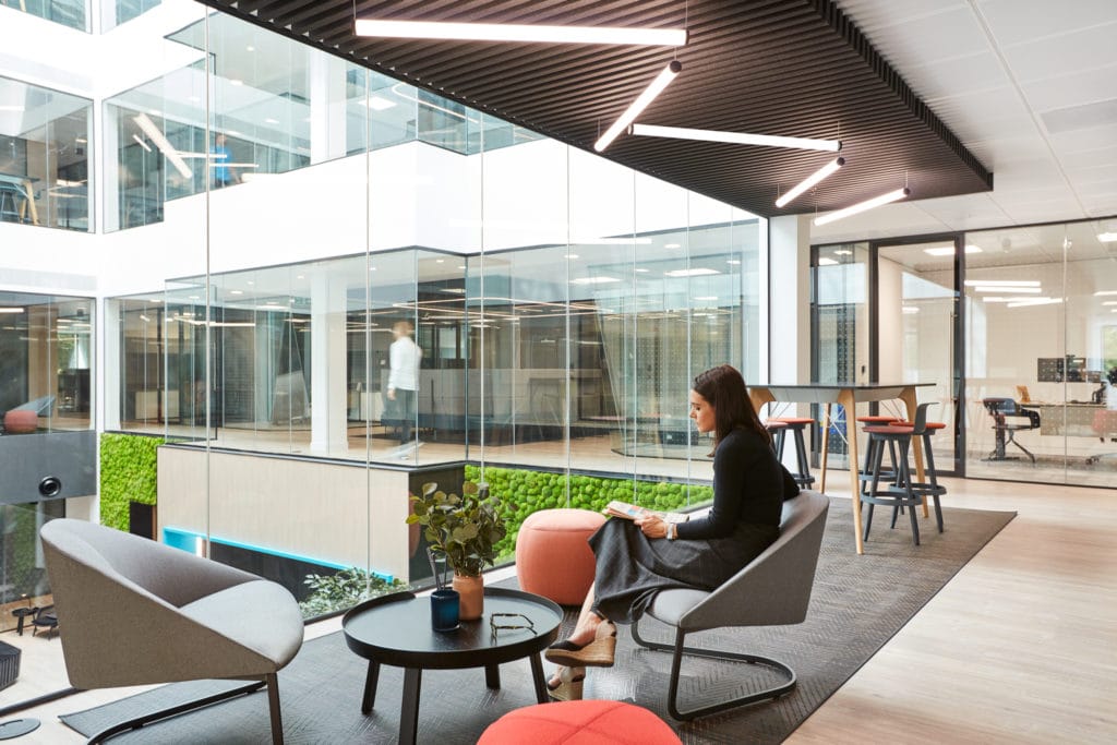 Open plan workplace seating area with a mixture of seating with person overlooking onto the ground floor through floor to ceiling windows