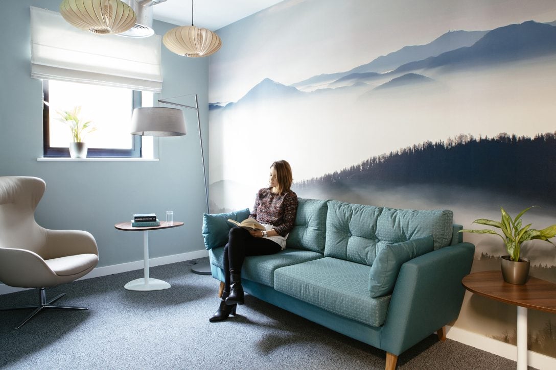 A female employee is shown sitting on a sofa reading inside a contemplation room in an office. The room is finished in soft tones of blue for a serene setting