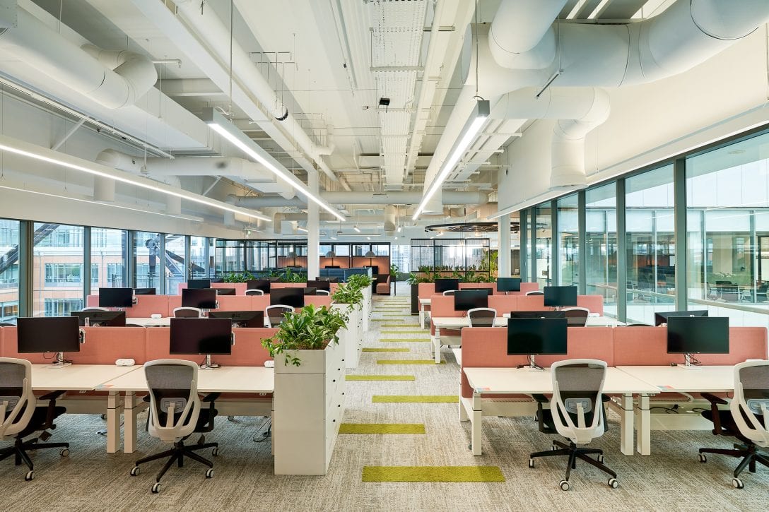 A colourful office is shown with many banks of desks which should be counted to calculate workplace densities, a useful and measurable workplace design metric. 