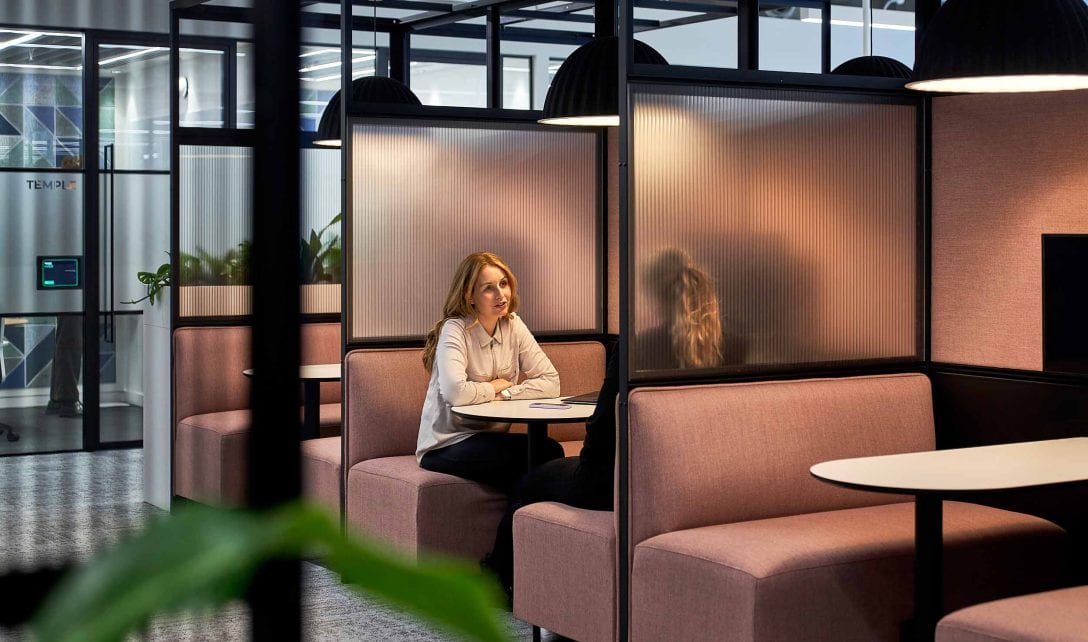 Two women are sat in a booth with dusty pink upholstery inside an AIS workplace design and build project.